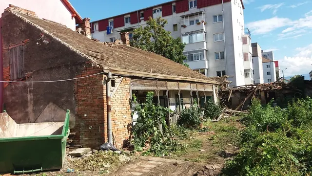 Régi, földszintes téglaház cseréptetővel és verandával a bontás előtt, mellette zöld sittkonténer, háttérben lakóépület