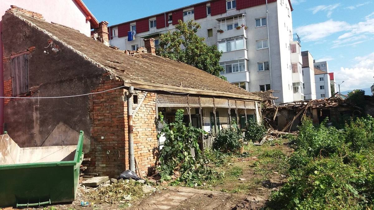 Régi, földszintes téglaház cseréptetővel és verandával a bontás előtt, mellette zöld sittkonténer, háttérben lakóépület